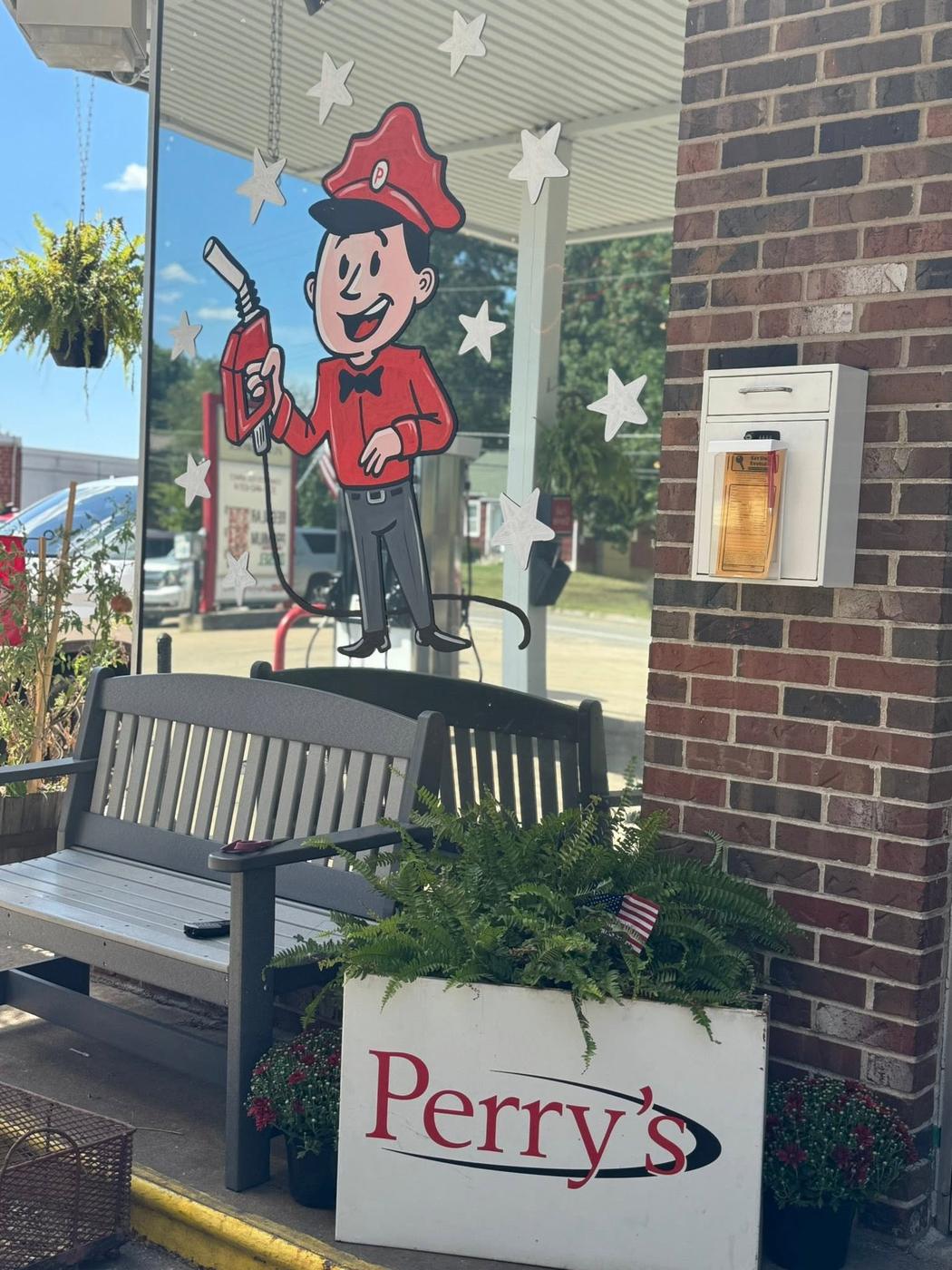A painted service attendant in a red cap holding a fuel nozzle on the station window, above a bench and a planter carrying the Perry's script logo