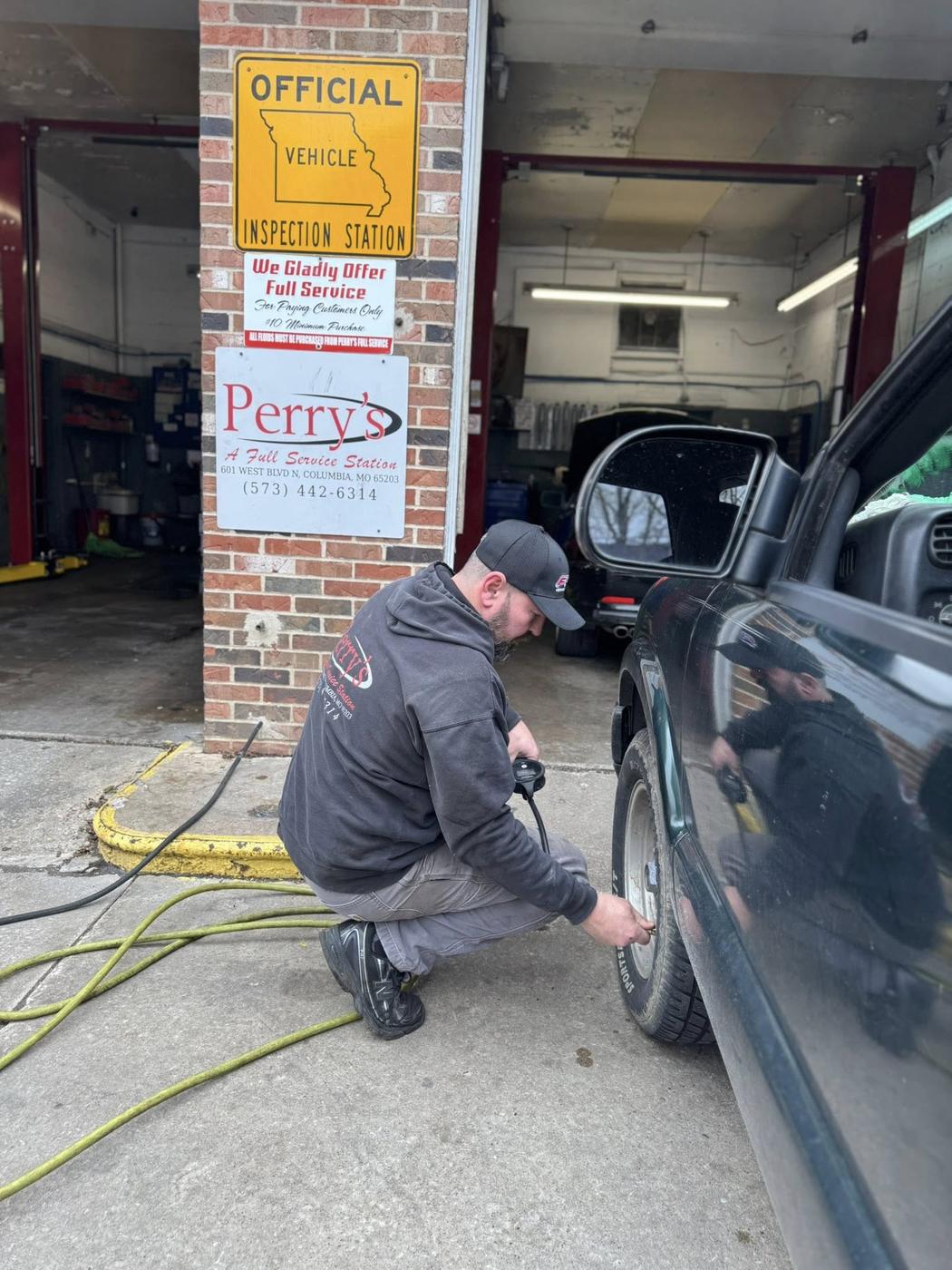 A technician kneeling at a car door inside the bay, with the Perry's Full Service Station sign on the pillar